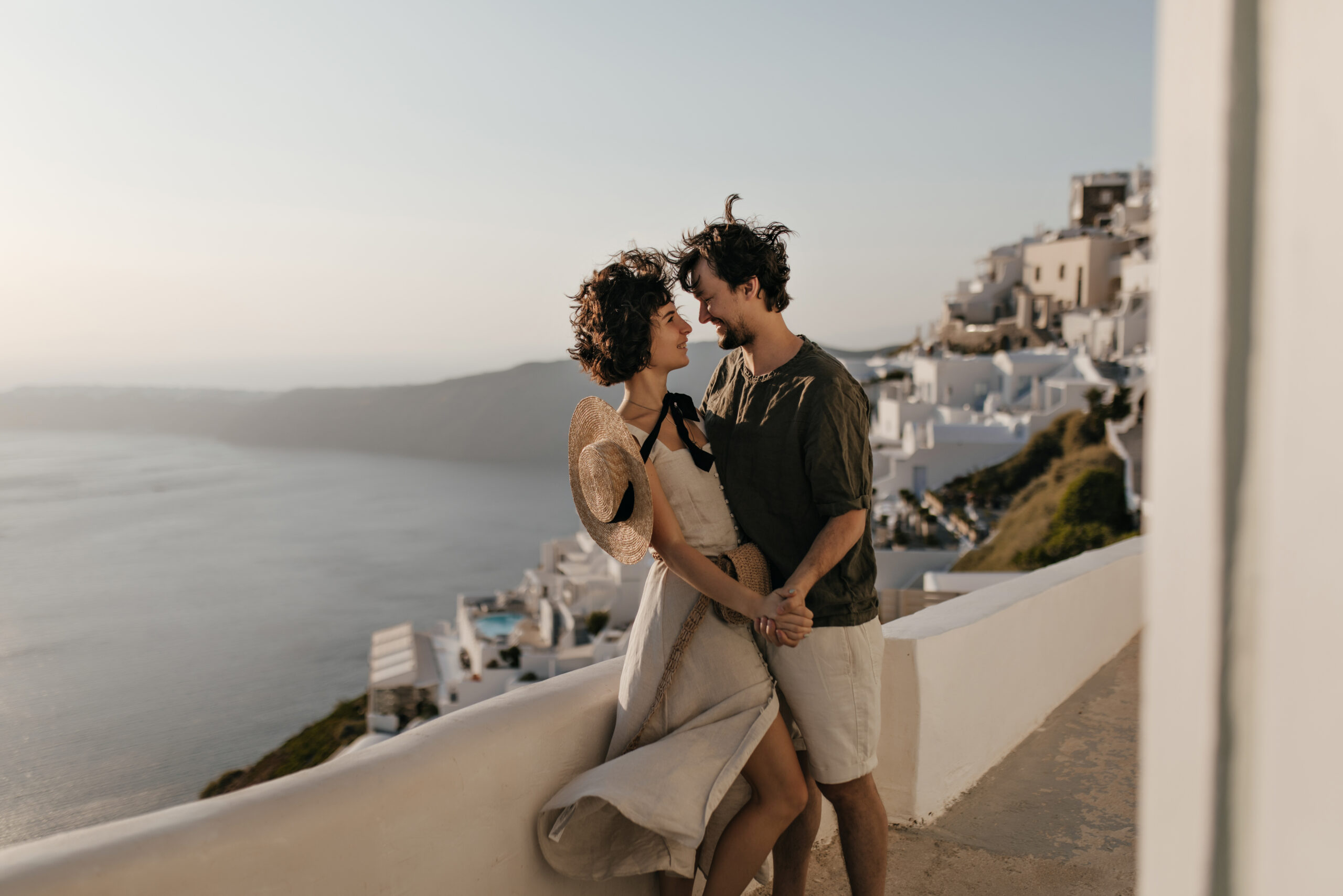 Portrait of couple on background of Greek city. Brunette lady in beige dress and man in stylish outfit looks at each other with love on sea background.